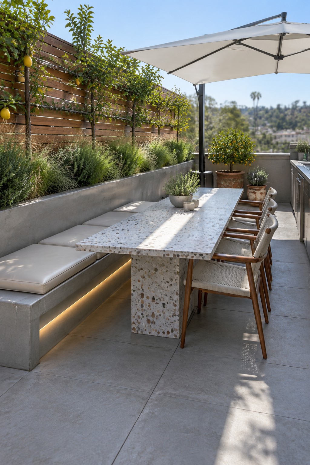 A roof terrace with a long terrazzo dining table, built-in concrete bench with cushions on one side, woven chairs on the other, and a raised planter wall with trees and shrubs behind the bench.