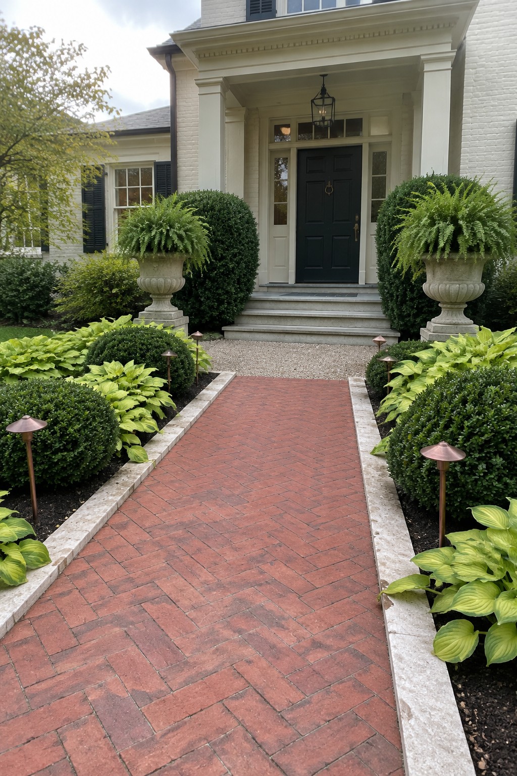 A red brick walkway bordered by hostas leads to the front steps of a white brick house.
