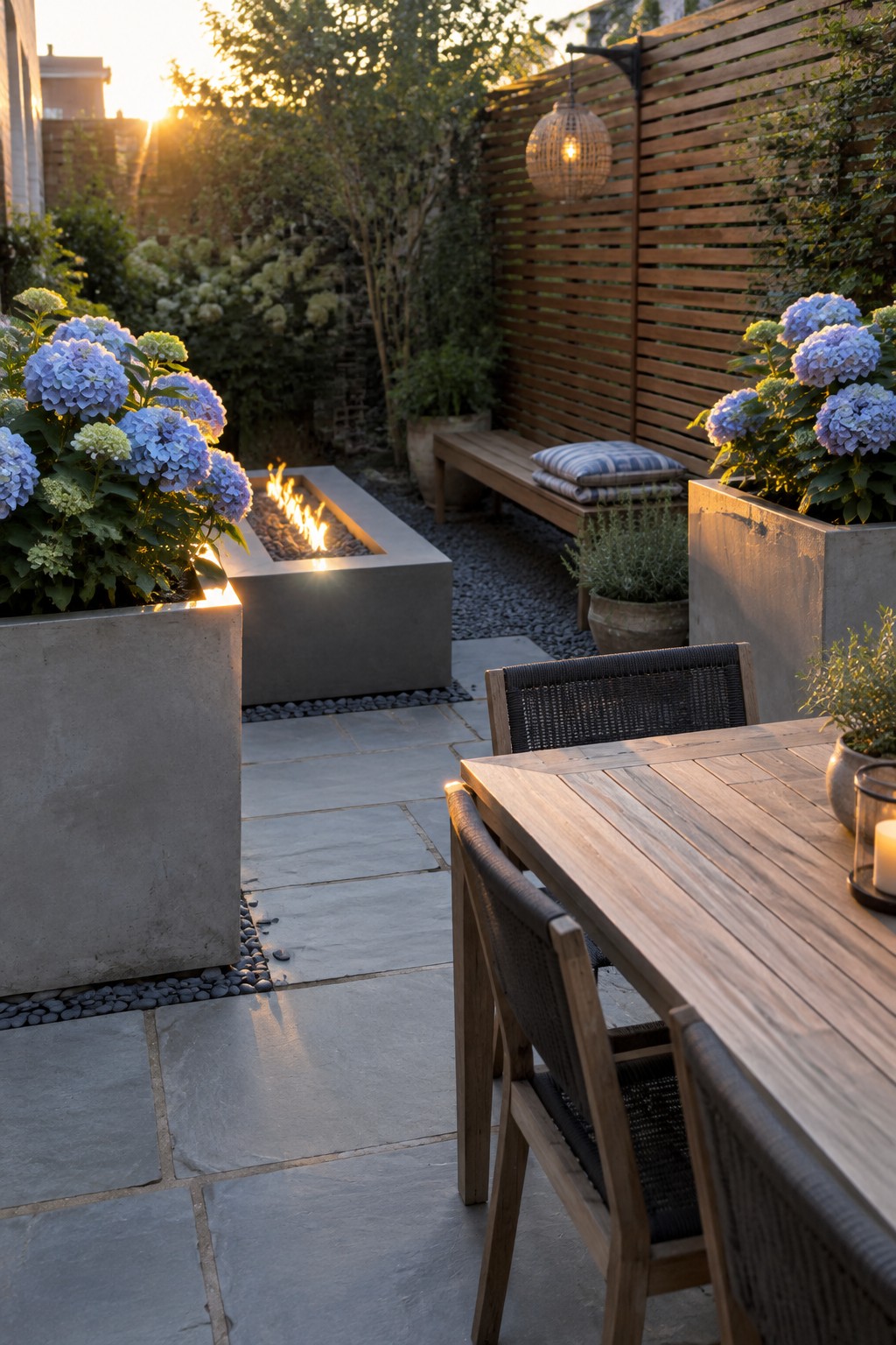 Blue hydrangeas in concrete planters beside a stone patio with a rectangular fire pit and wooden dining furniture.