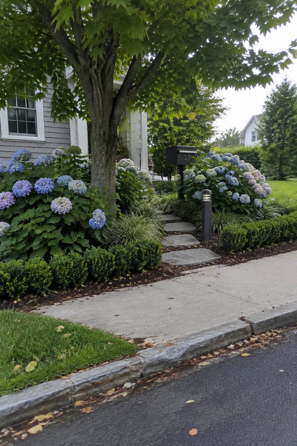 Stepping stones wind through hydrangeas and ornamental grasses beneath a mature tree beside a house.