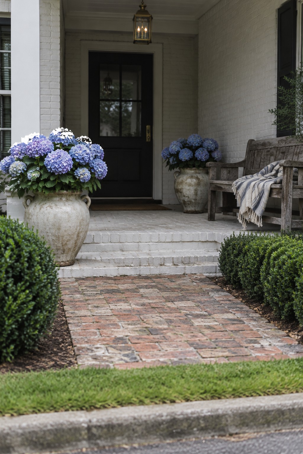 Blue and purple hydrangeas bloom in large ceramic pots beside a brick porch path and wooden bench.