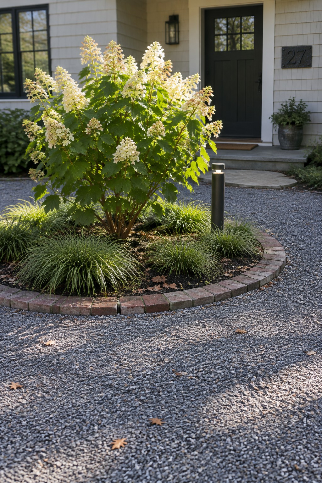 White hydrangeas and ornamental grasses grow in a curved garden bed edged with bricks beside a gravel driveway.