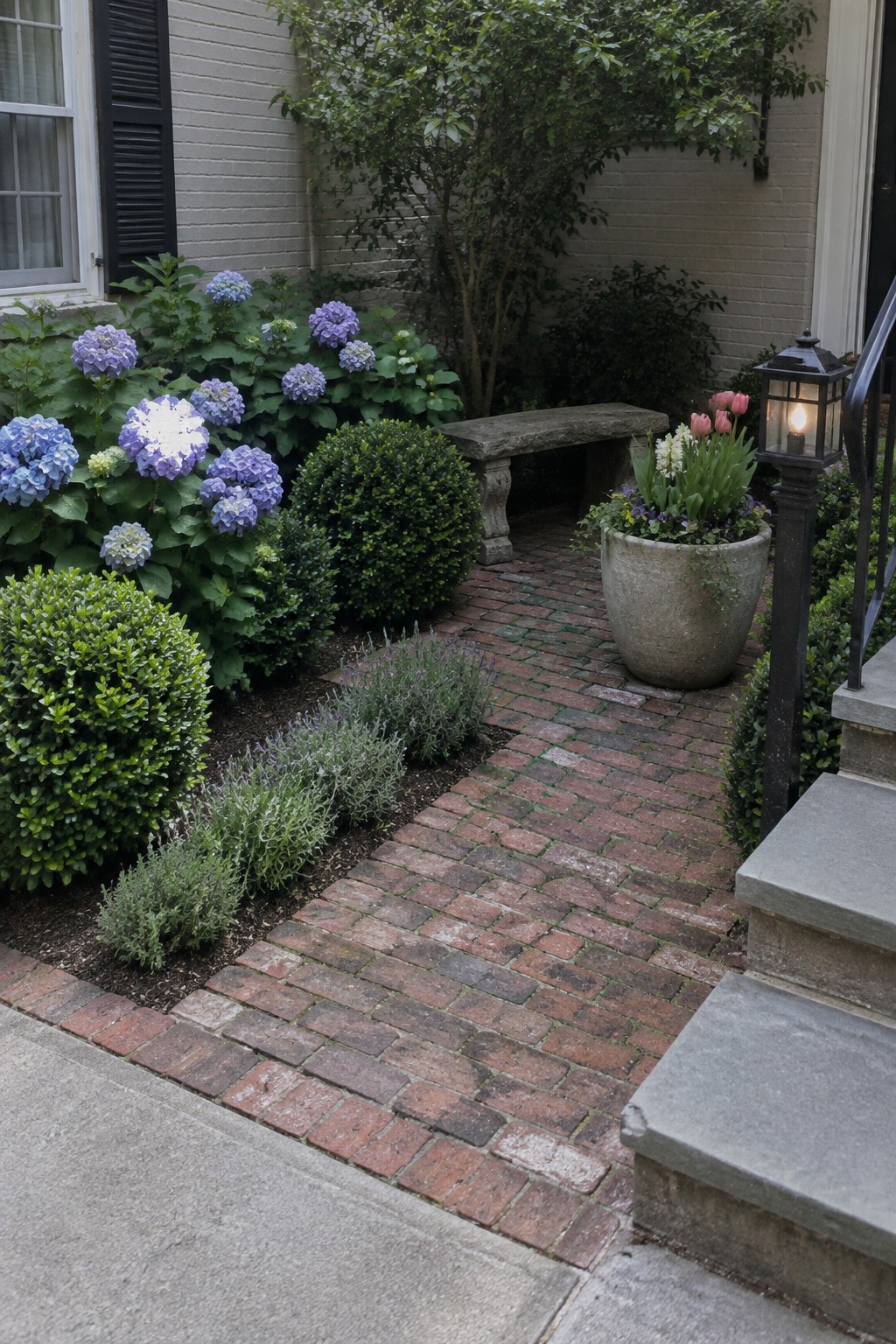 A brick walkway passes hydrangeas and clipped shrubs beside a light-colored brick house.