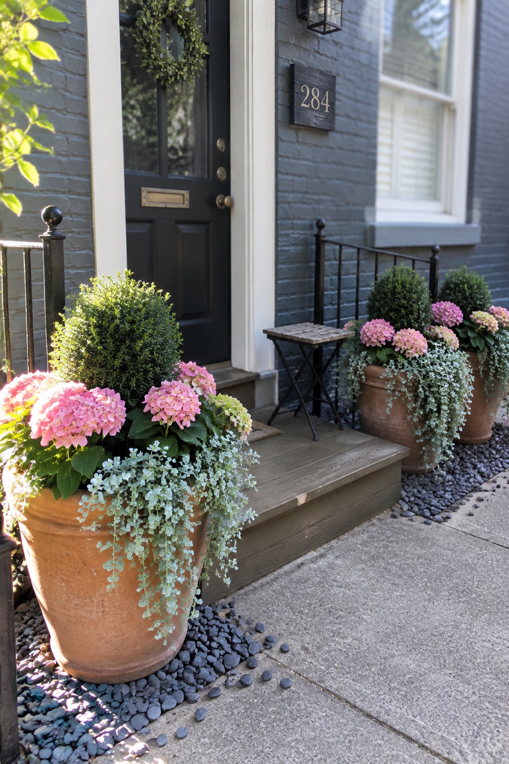 Large terracotta pots filled with pink hydrangeas and trailing plants sit beside the steps of a brick house.