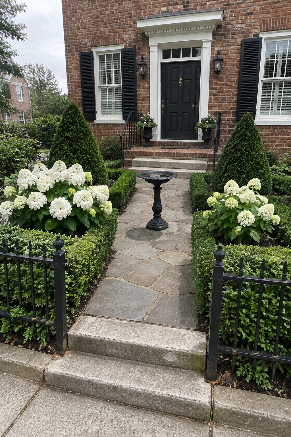 White hydrangeas and clipped low hedges border a stone walkway leading to the steps of a brick house.