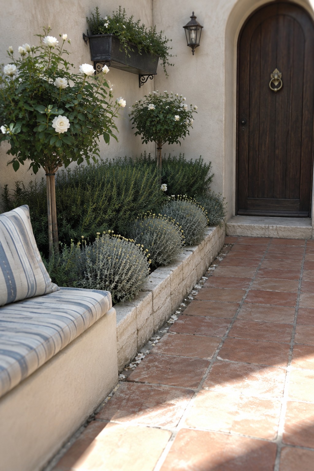 White rose bushes and low plants grow in a raised stone bed beside a terracotta-tiled walkway and a dark wooden front door.