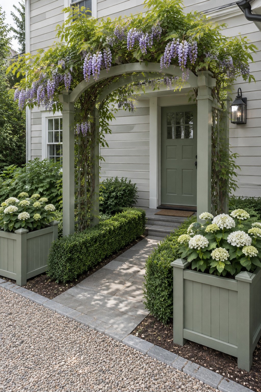 A wisteria-covered garden arch stands over a walkway leading to a green front door, with white hydrangeas in raised planters.