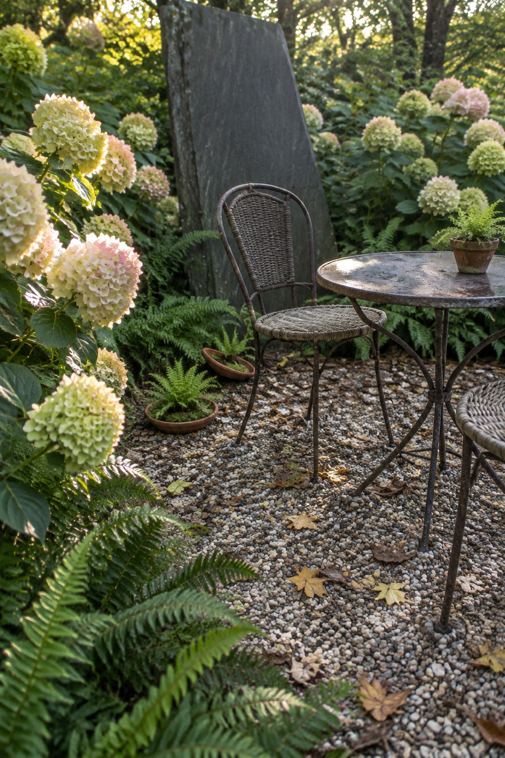 A metal table and woven chairs sit on a gravel clearing among ferns and flowering hydrangeas.