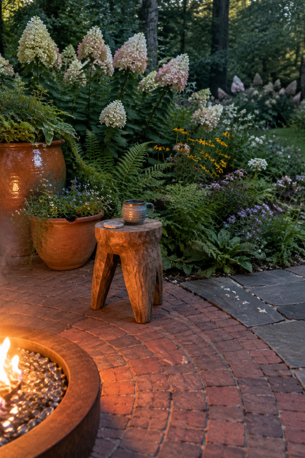 A brick patio with a round fire pit beside terracotta pots, a wooden stool, hydrangeas, and ferns.