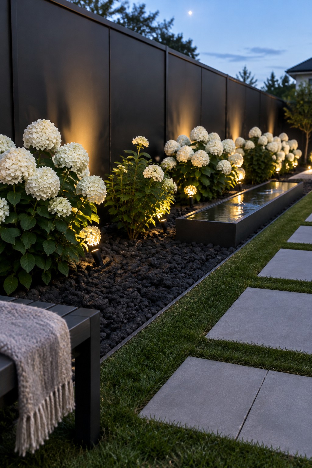 White hydrangeas grow along a dark fence beside a long water feature, with small garden lights placed in the planting bed.