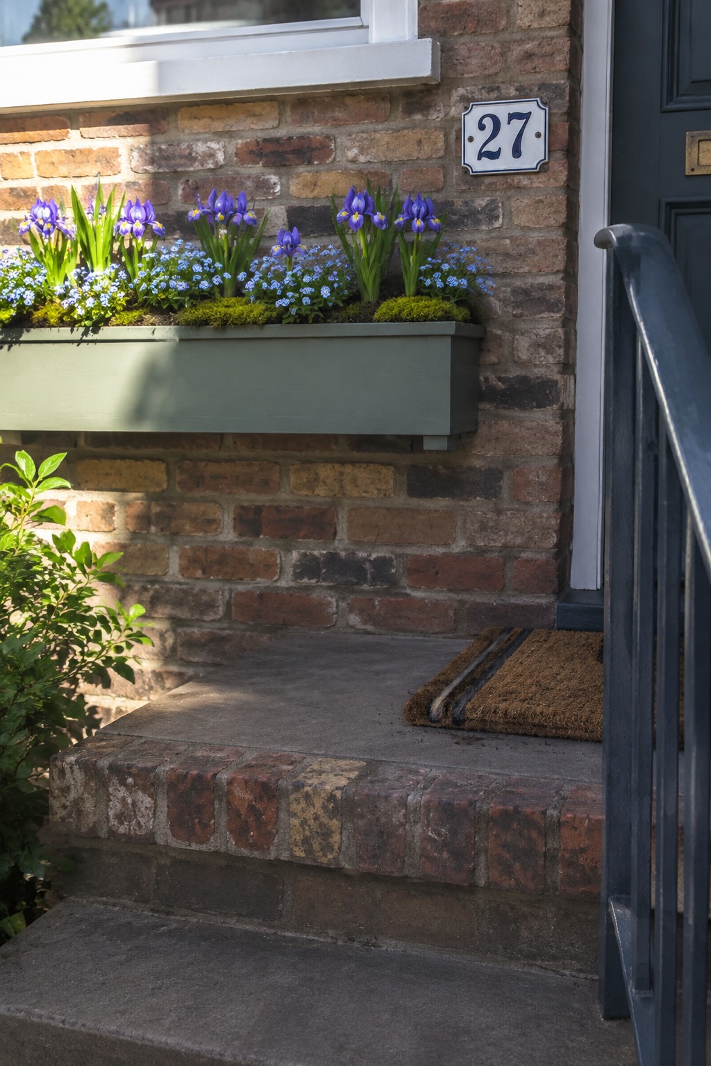 A long gray window box filled with purple irises and small blue flowers sits against a brick wall beside a front door marked 27.