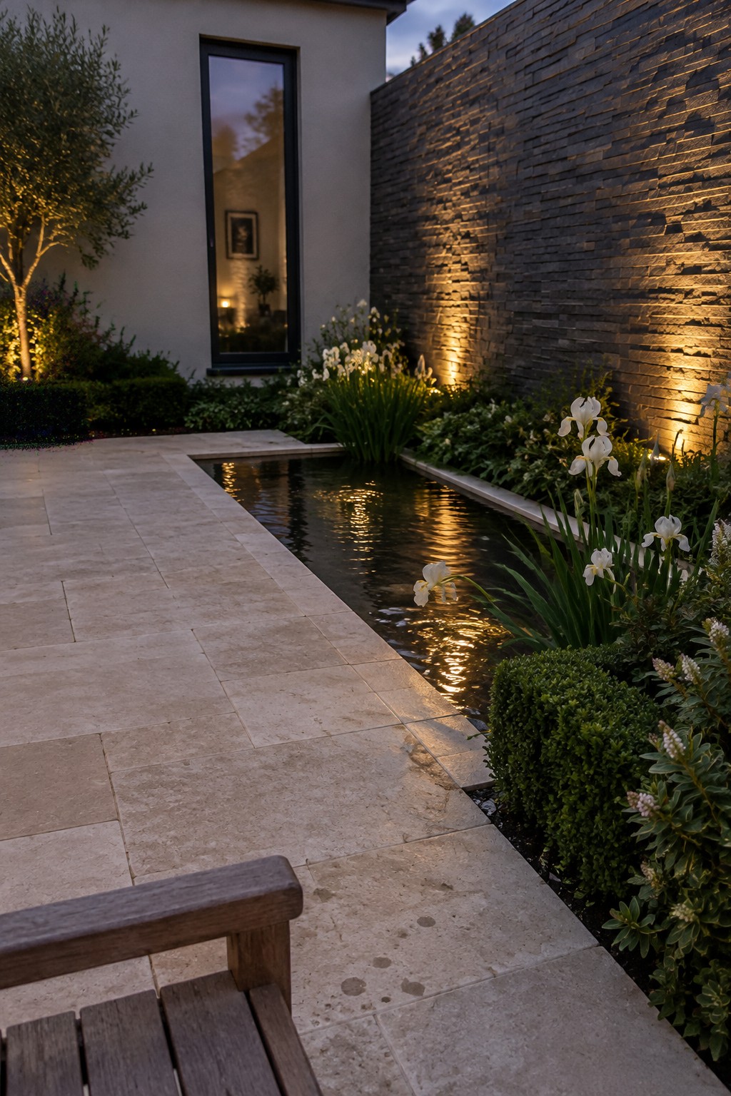 White irises grow beside a narrow reflecting pool bordered by pale stone paving and a dark stacked-stone wall.