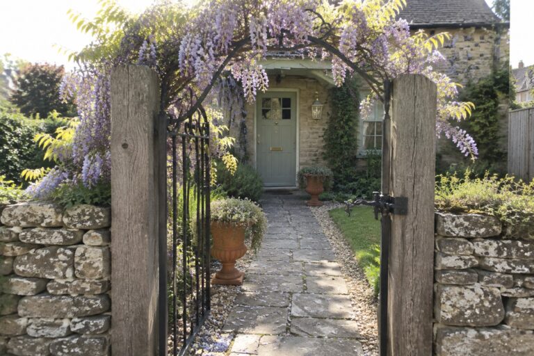 19 Wisteria Entrance Ideas for Gates and Walkways A stone path passes through an open iron gate beneath a wooden arch covered in purple wisteria.