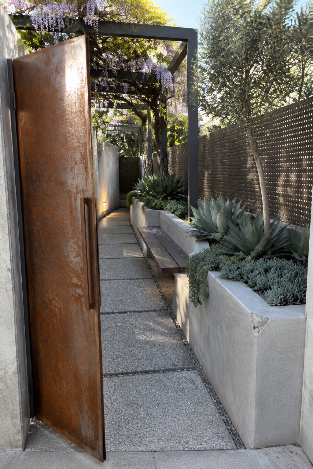 An open rusted metal gate leads to a narrow garden walkway beneath a dark pergola covered in purple wisteria.