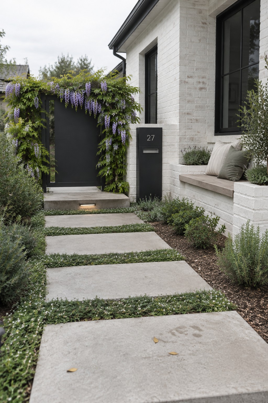 A concrete stepping-stone path leads to a black garden gate covered in flowering wisteria beside a white brick house.