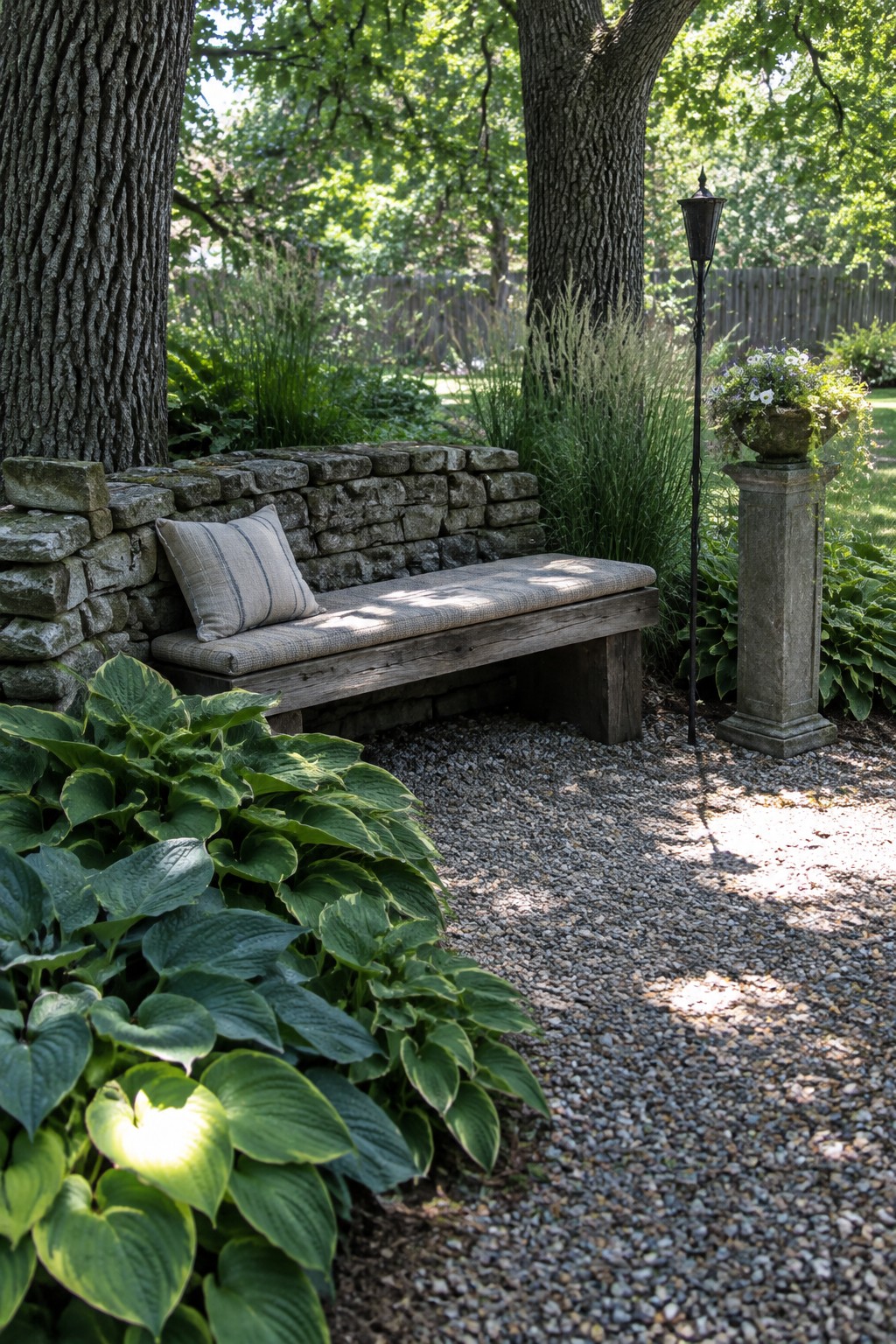A wooden bench with a cushion sits beside a low stone wall and gravel path among hostas and mature trees.