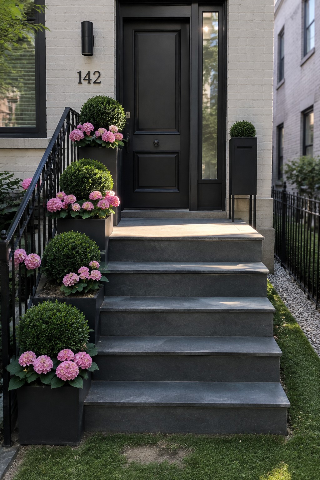 Black planters filled with rounded boxwoods and pink hydrangeas line dark stone steps leading to a black front door.
