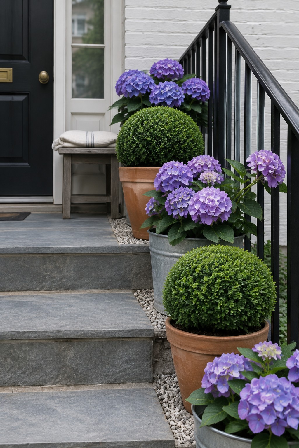 Purple hydrangeas and clipped boxwood shrubs in pots line gray stone steps beside a black front door.