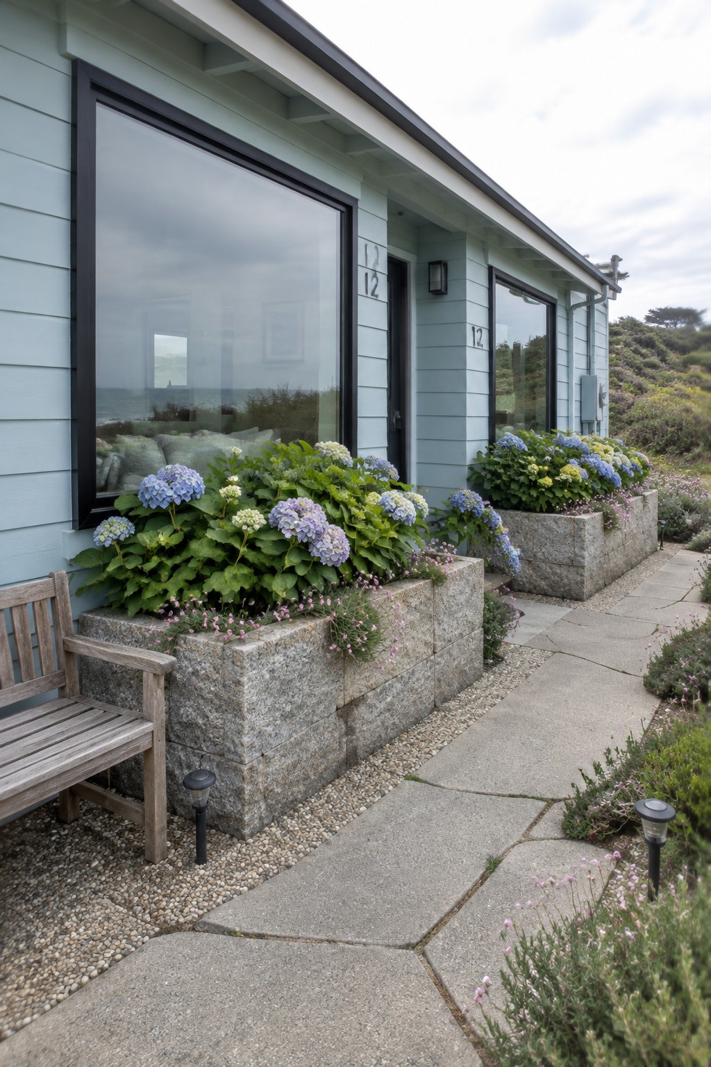 Blue and pink hydrangeas grow in raised stone beds beside a concrete walkway along a pale blue house.