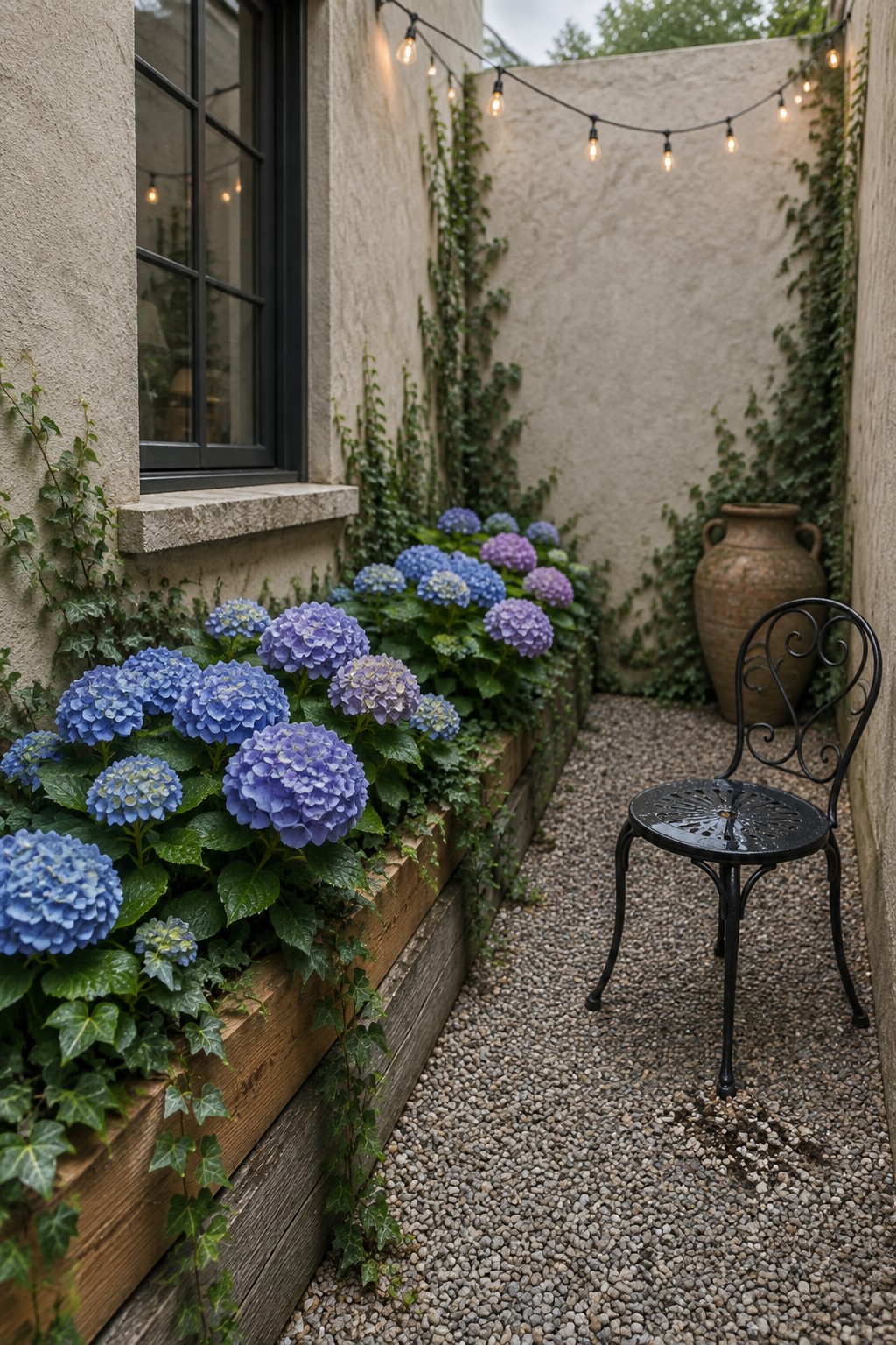 Blue and purple hydrangeas grow in a raised timber bed beneath a window beside a narrow gravel path, with a metal chair nearby.