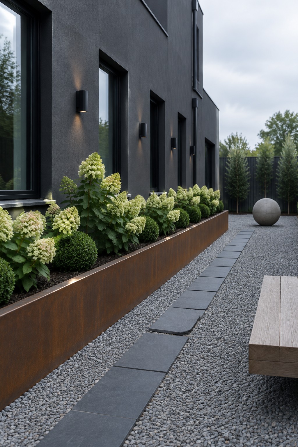 White hydrangeas and round shrubs grow in a rust-colored raised planter beside a dark house, gravel path, and rectangular stepping stones.