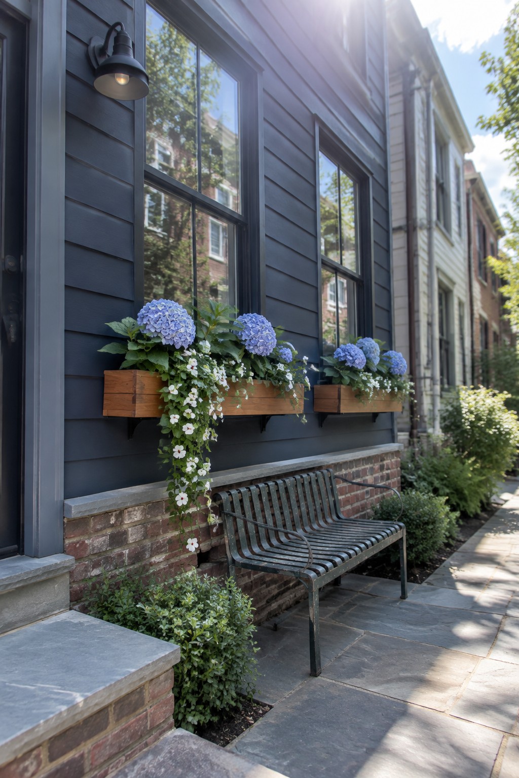 Wooden window boxes filled with blue hydrangeas and trailing white flowers hang beneath windows on a dark-sided house.
