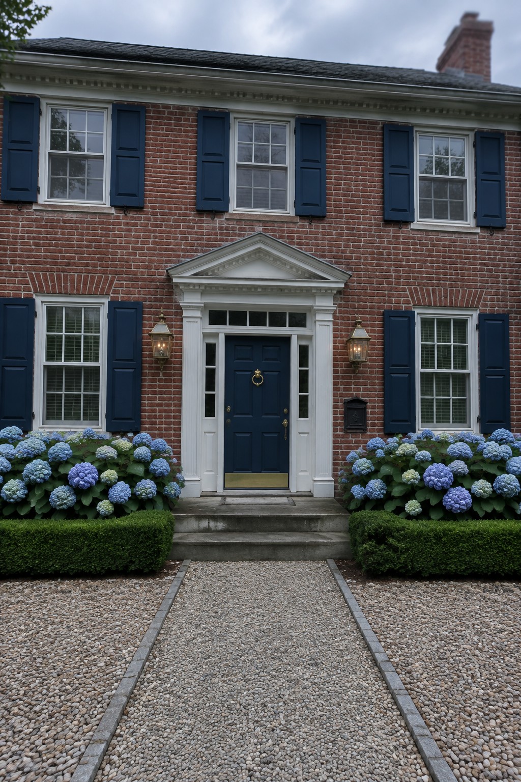 Blue and purple hydrangeas grow in beds beside the gravel path leading to a brick house’s front entrance.