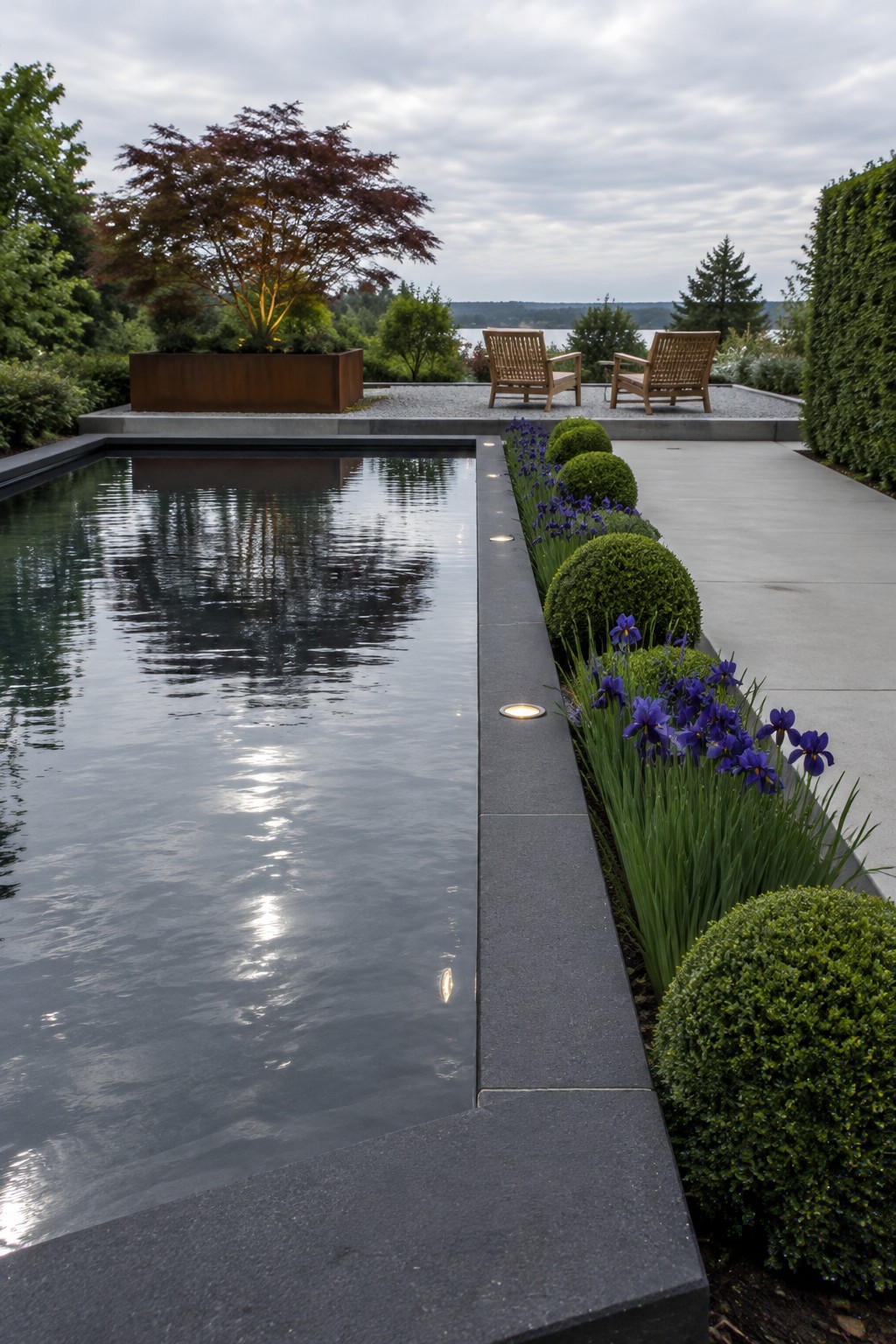 Purple irises and rounded shrubs grow beside a rectangular reflecting pool with a paved walkway.