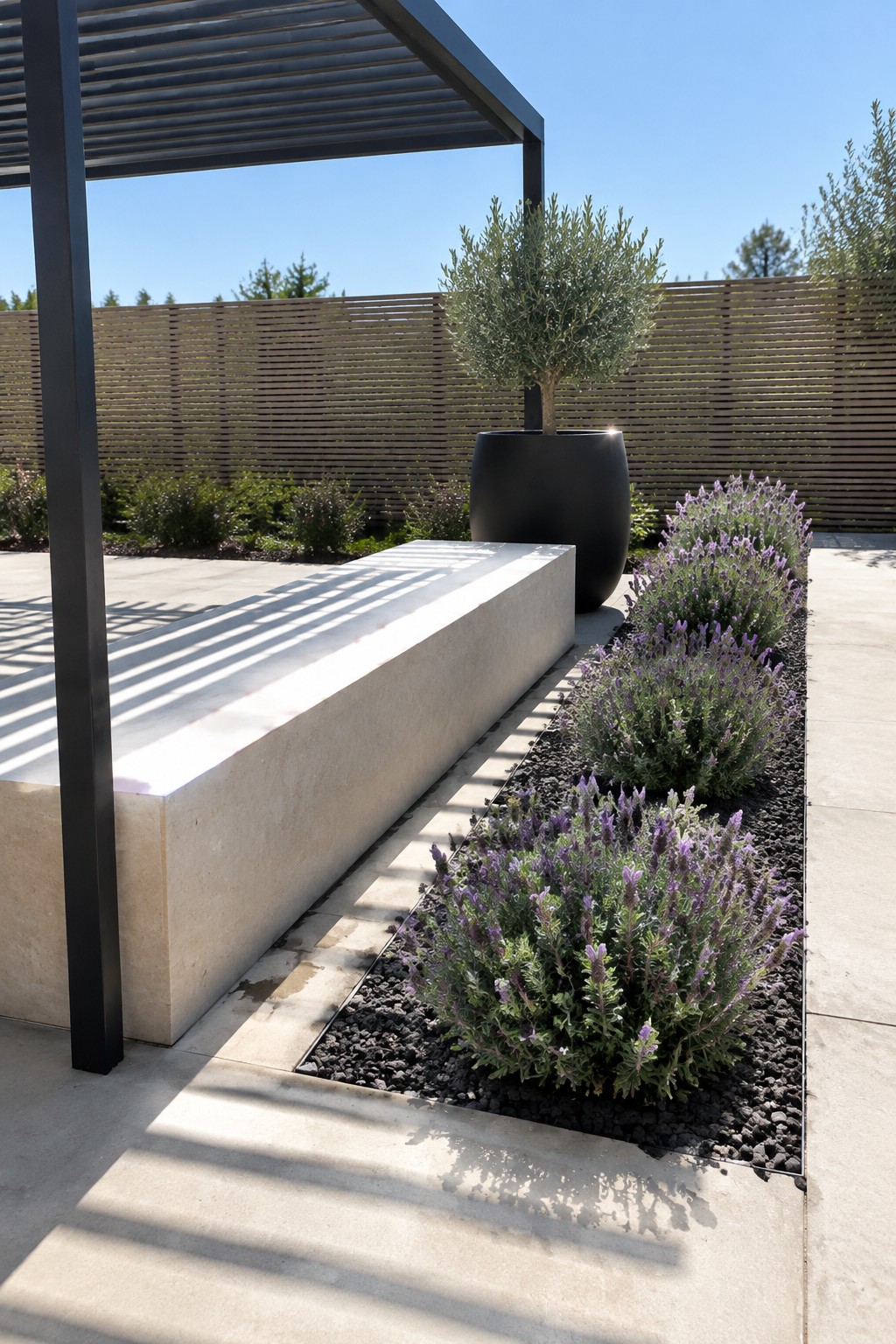 A narrow bed of purple lavender grows in dark gravel beside a pale stone bench on a paved patio.