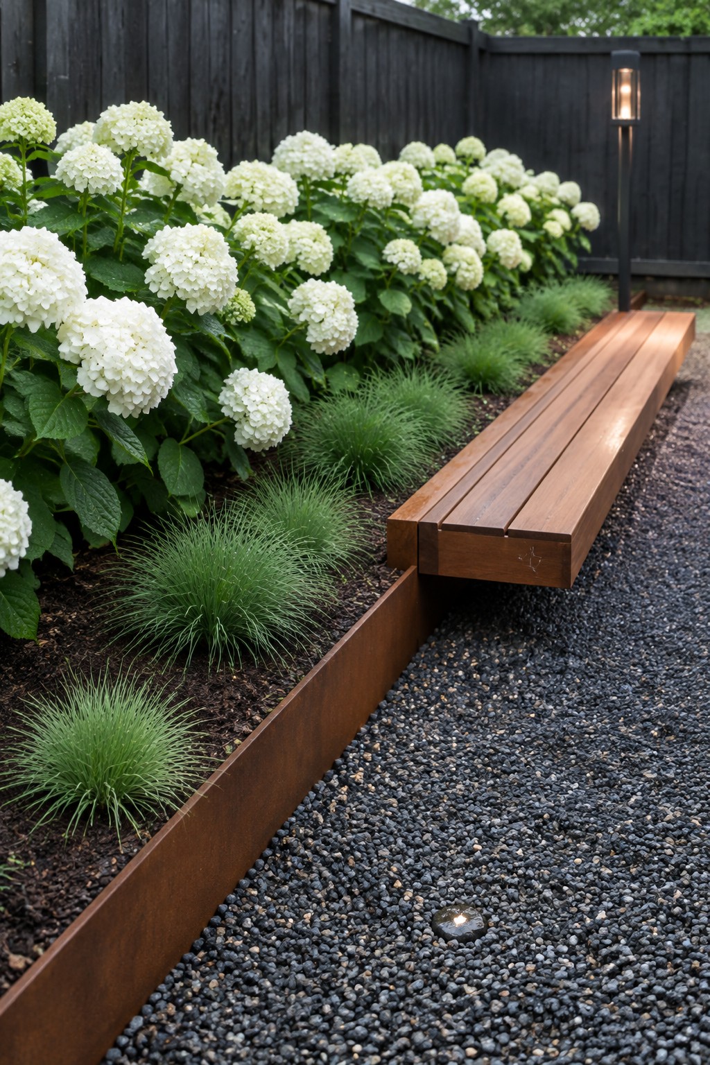 White hydrangeas and ornamental grasses grow beside a gravel path with a long wooden bench built into the raised bed edge.