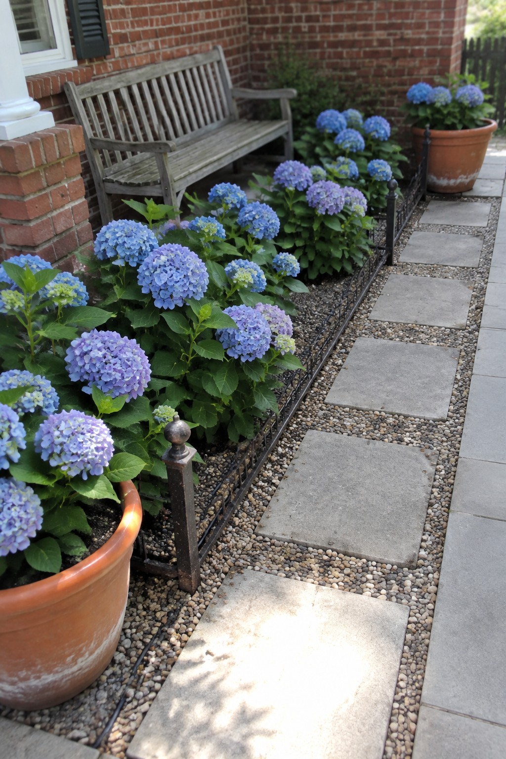 Blue and purple hydrangeas grow beside a gravel path lined with stone pavers and low metal edging.