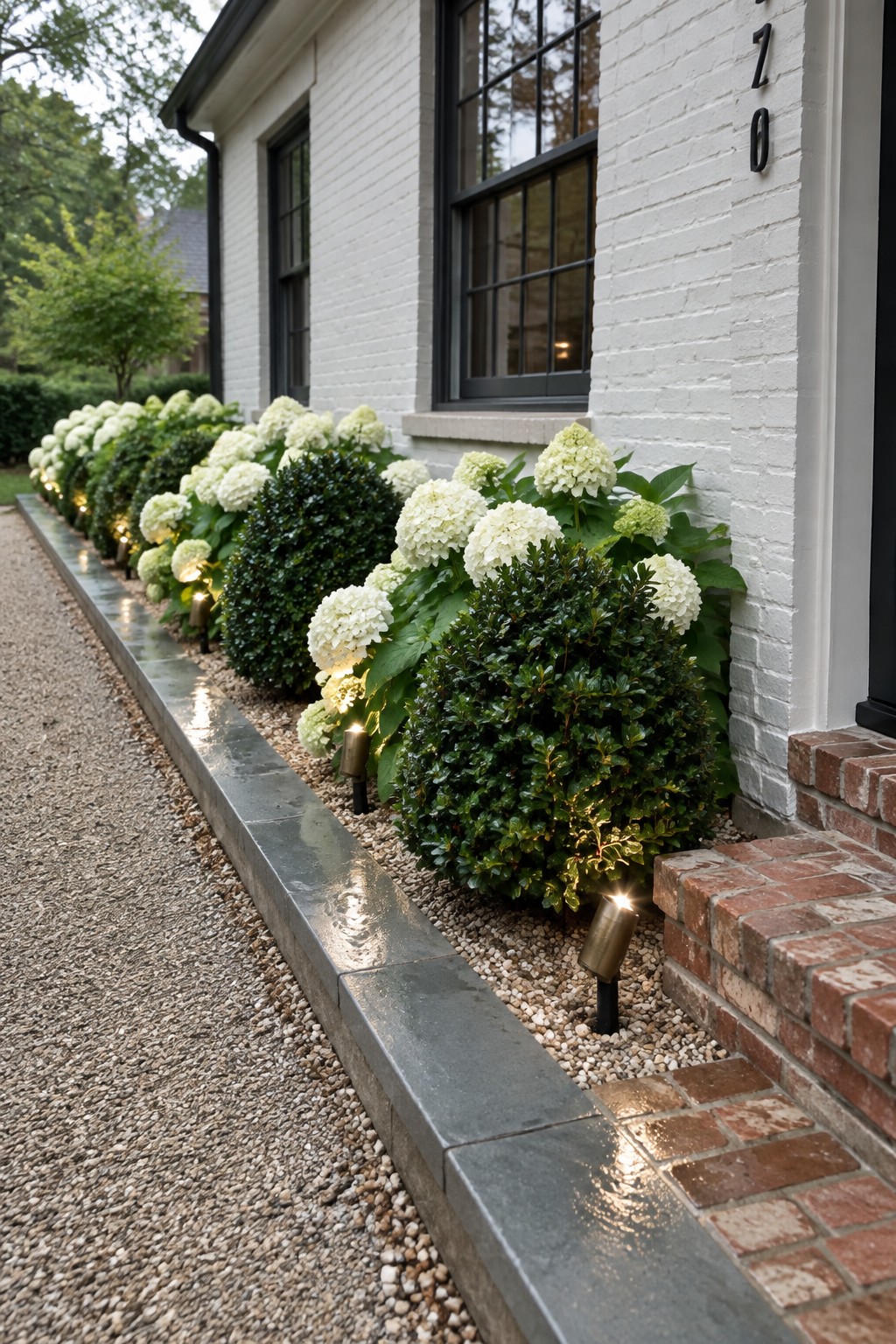 White hydrangeas and rounded evergreen shrubs repeat along a gravel garden bed beside a white brick house.