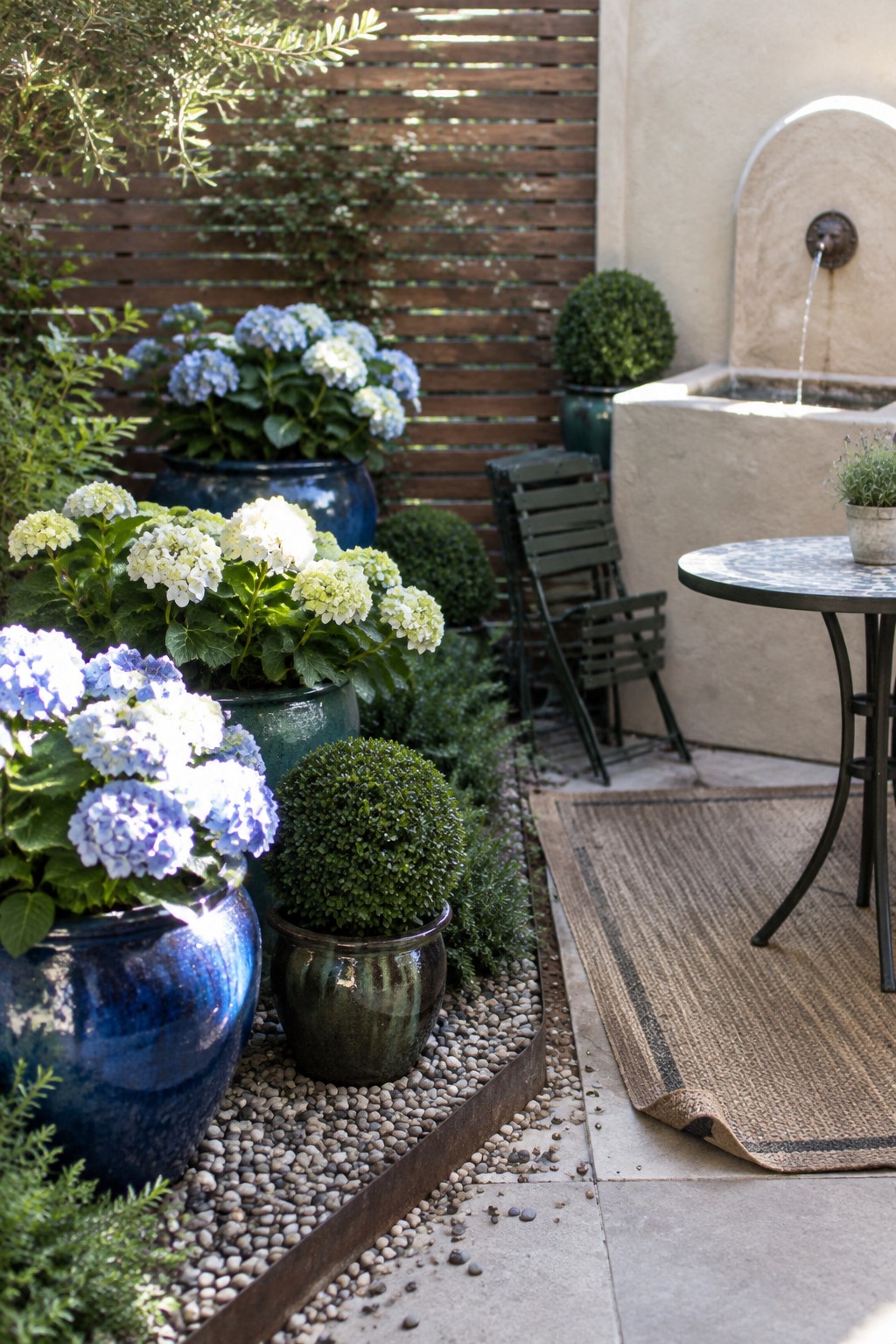 Blue and white hydrangeas grow in large ceramic pots beside a small patio table and a wall fountain.