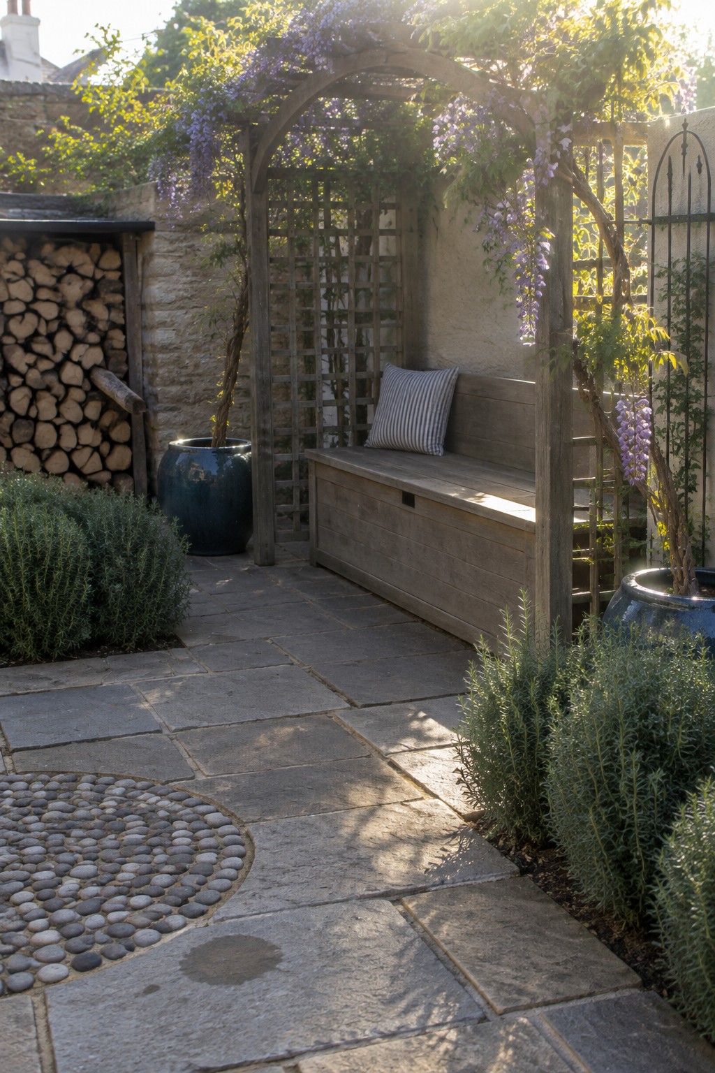 A wooden bench beneath a wisteria-covered trellis in a small stone-paved garden.