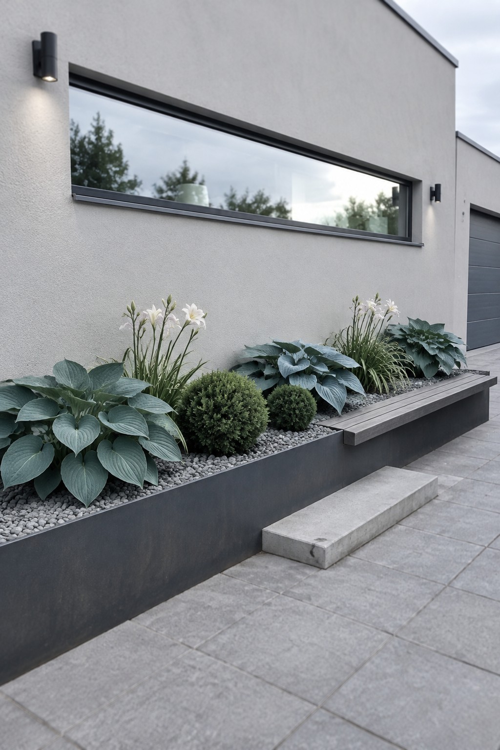 A raised garden bed with blue-green hostas, small shrubs, and white flowers runs along a light stucco wall beneath a horizontal window.