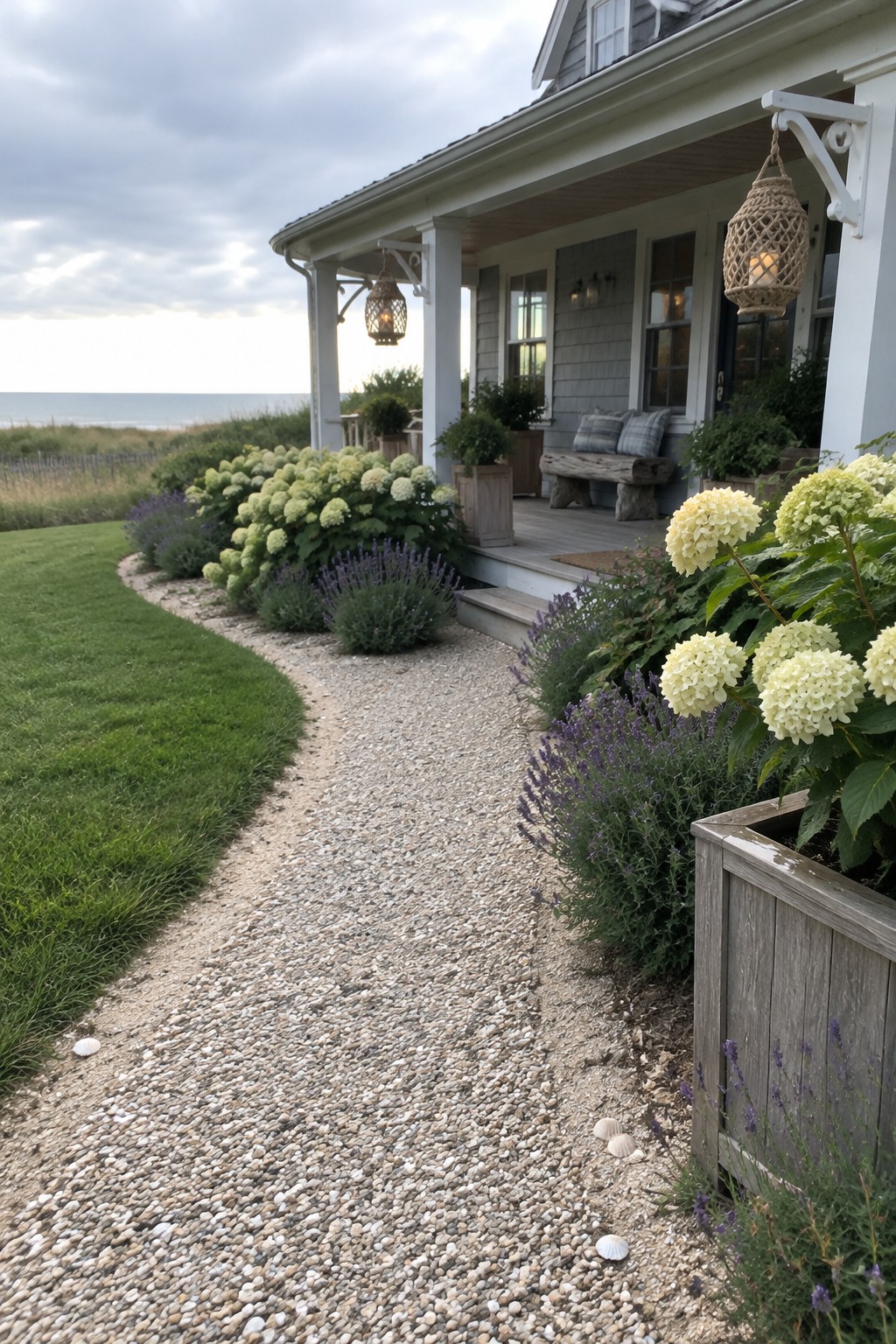 A curved pebble walkway passes hydrangeas and purple flowers beside a coastal home’s porch.