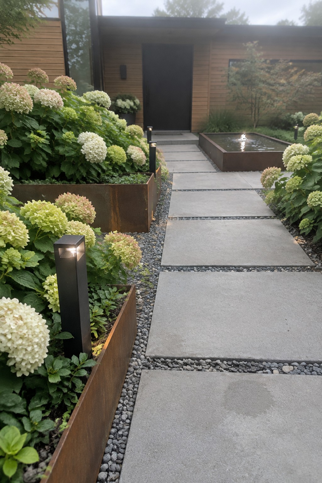 Large concrete stepping pads run between raised metal beds planted with hydrangeas, leading to a dark entry door.