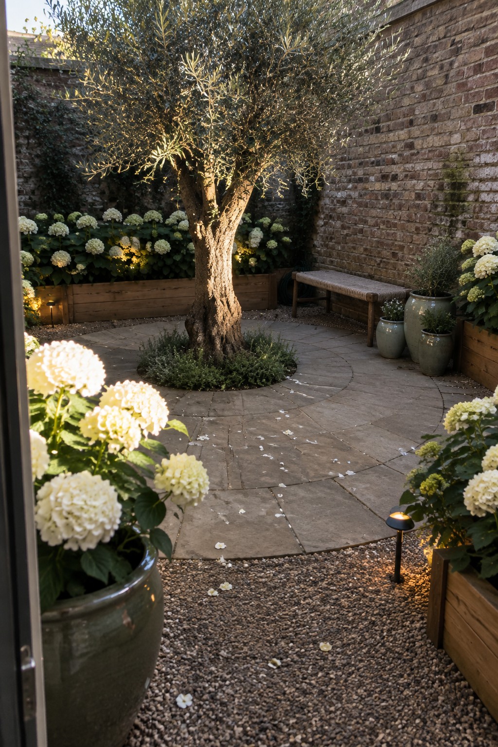A mature tree stands in a circular paved courtyard surrounded by white hydrangeas, with a wooden bench and potted plants beside brick walls.