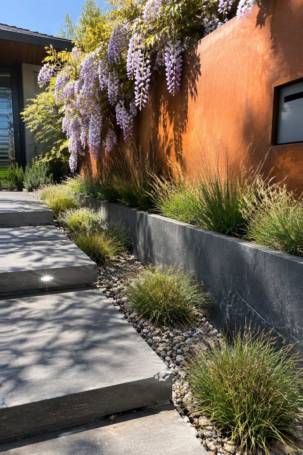 Purple wisteria cascades over a rust-colored wall above a raised stone bed beside a paved walkway.