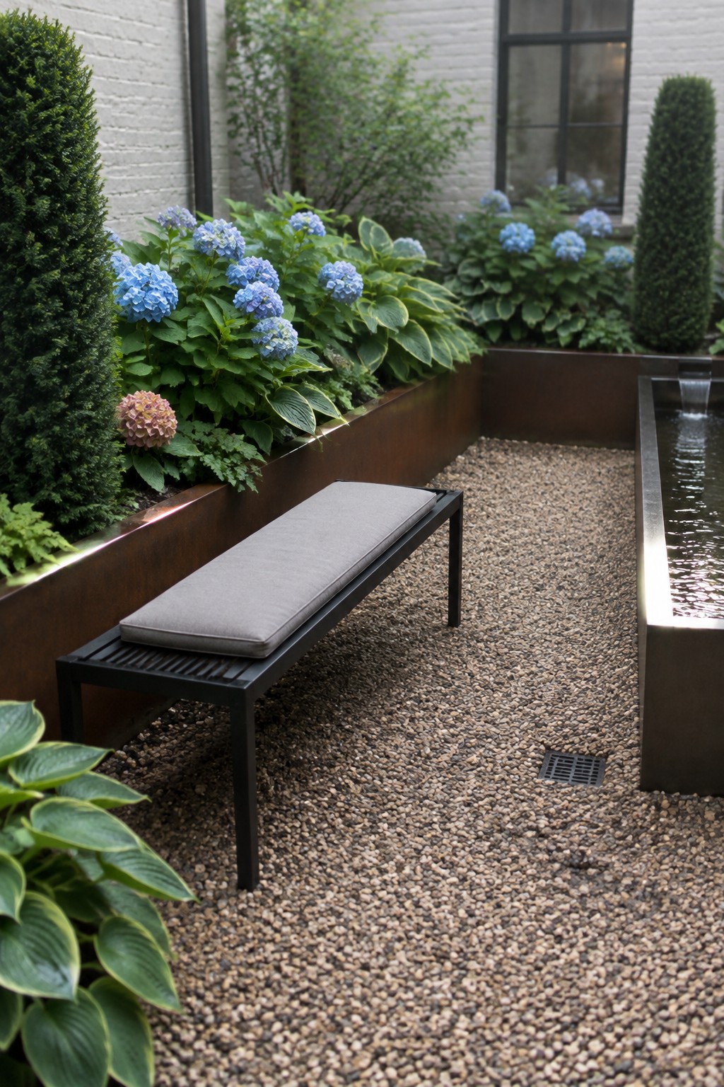 A raised rust-colored garden bed filled with hostas and blue hydrangeas beside a gravel courtyard, bench, and narrow water feature.