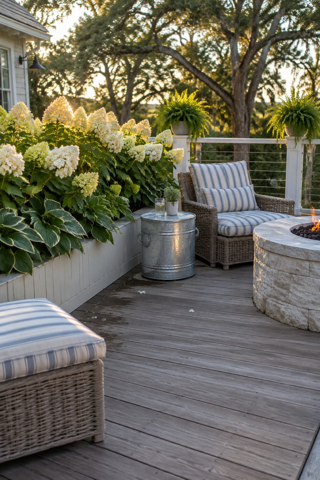 White hydrangeas and hostas fill a raised bed beside a wooden deck with wicker seating and a stone fire pit.