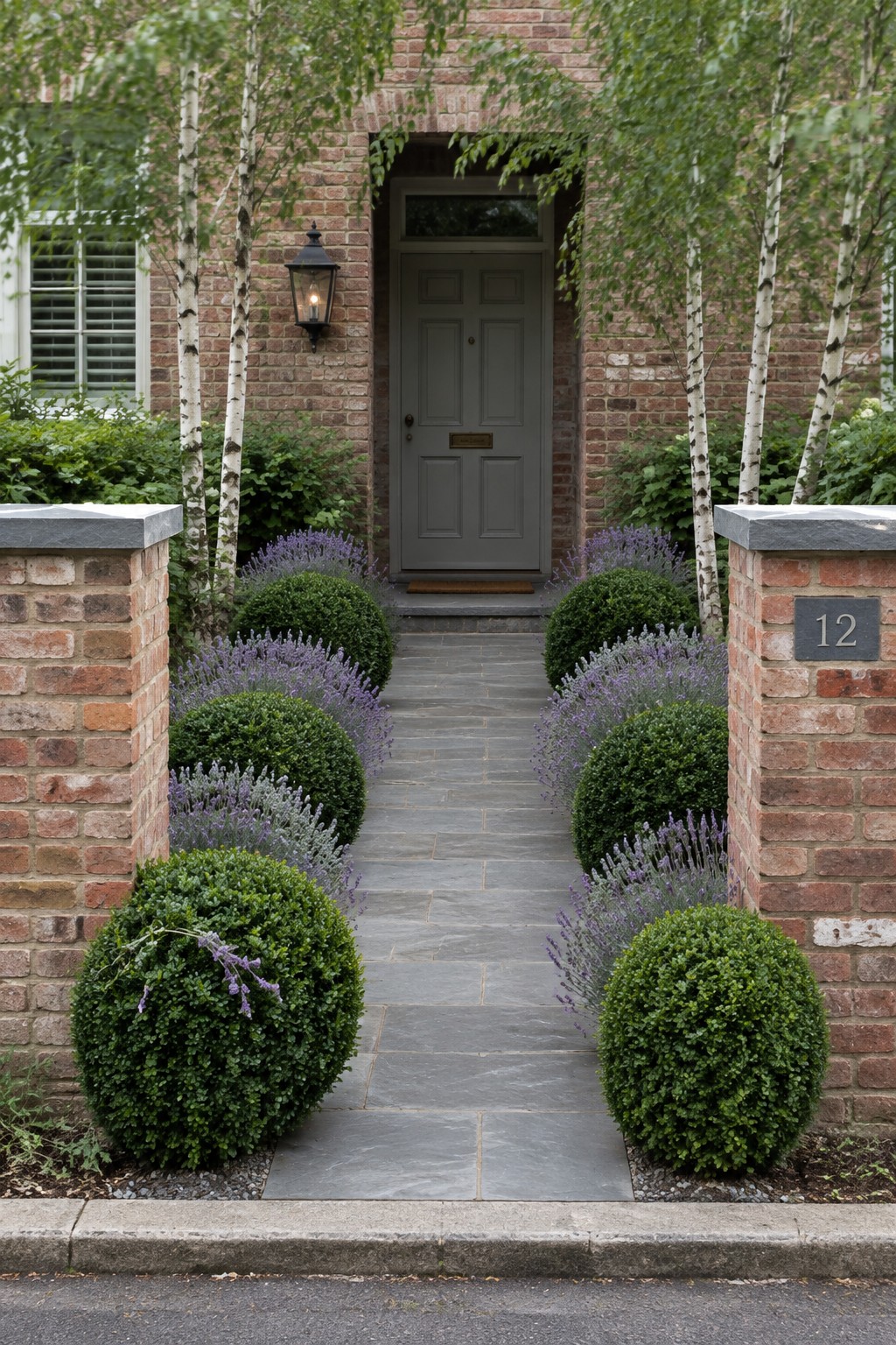 A straight gray stone path leads between rounded boxwood shrubs and purple lavender to a dark front door, with brick pillars on either side.