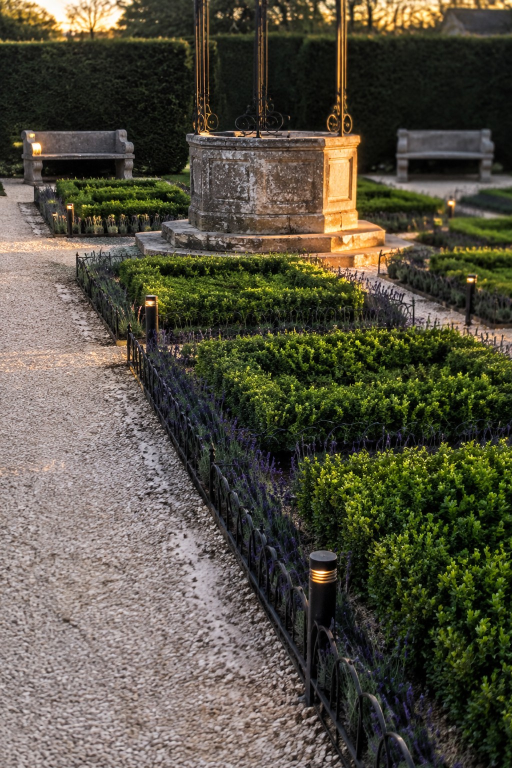 Clipped boxwood beds edged with lavender and low black fencing beside a gravel path, with a stone garden well and benches in the background.